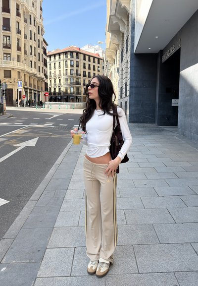 Joven mujer con cabello largo y oscuro, lleva gafas de sol, top blanco, pantalones deportivos beige, sostiene una bebida, de pie en una acera de la ciudad cerca de edificios altos.