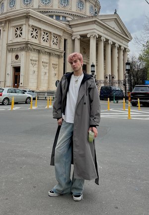 Persona joven con cabello rosa que lleva un abrigo largo gris, camisa blanca y jeans anchos, sostiene una bebida verde, de pie frente a un gran edificio neoclásico.