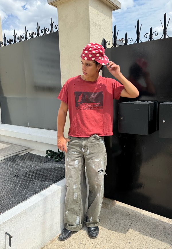 Jeune homme portant une casquette rouge à pois à l'envers, un t-shirt rouge et un pantalon gris déchiré, debout près d'une porte noire dans un cadre extérieur ensoleillé.