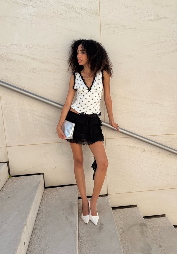Vrouw met krullend haar staat op de trap met een zilveren clutch in haar hand, draagt een wit top met stippen, een zwarte rok met ruches en witte hoge hakken, tegen een beige muur.