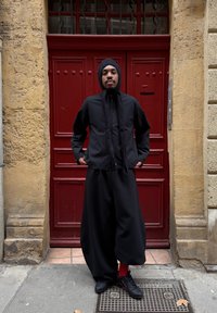 Svart jakke med glidelås og lange skjerf-lignende detaljer, kombinert med løstsittende svarte bukser og svarte joggesko. Rødt bakgrunn gir kontrast.