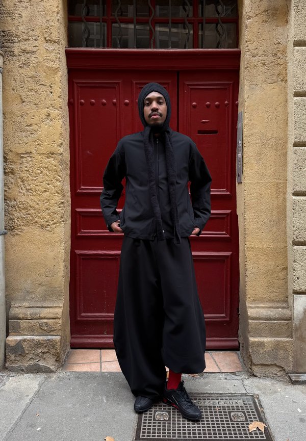 Juoda striukė su užtrauktuku ir ilgais šaliką primenančiais akcentais, derinama su laisvai krentančiomis juodomis kelnėmis ir juodais sportbačiais. Raudonas fonas suteikia kontrastą.