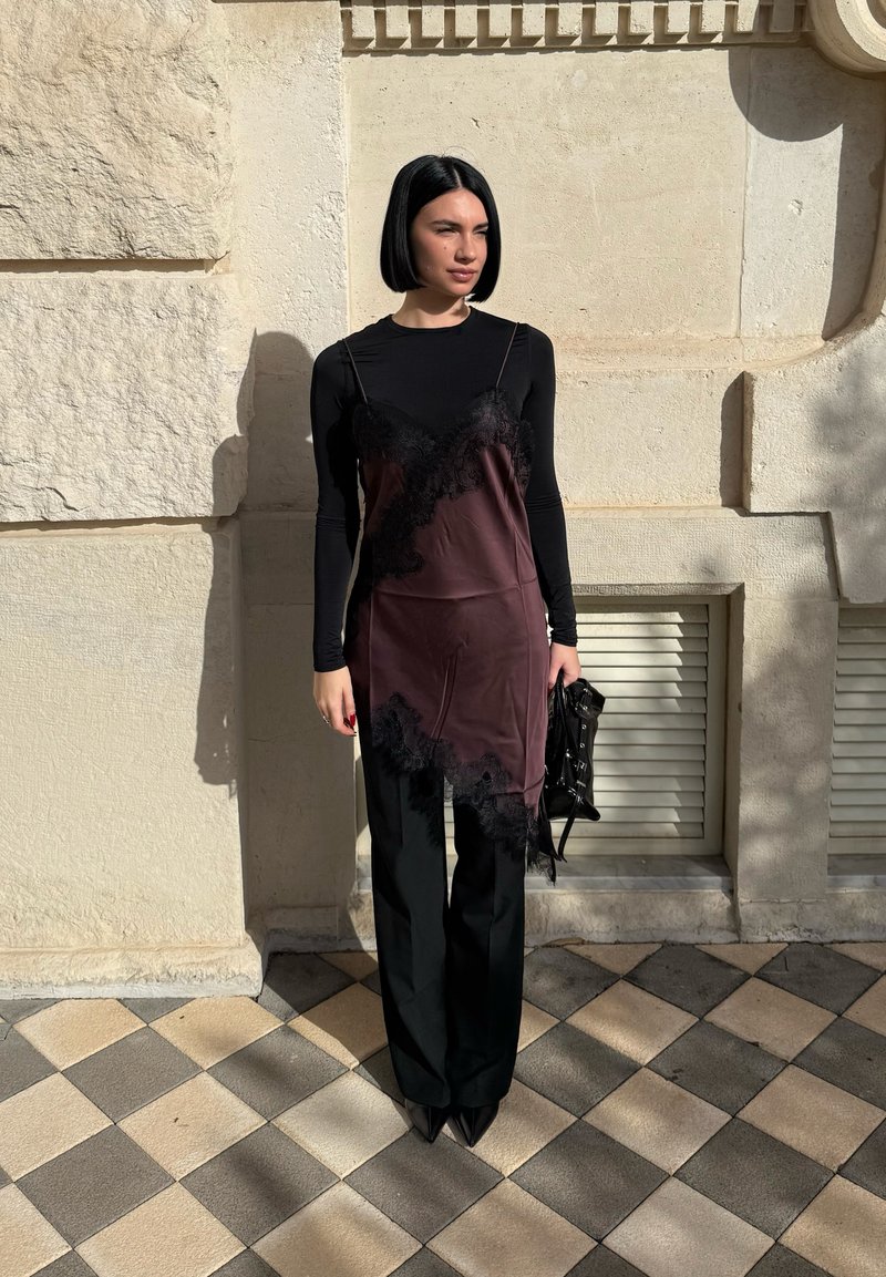 Femme avec coupe au carré noire portant un haut noir à manches longues, une robe portefeuille en dentelle violette, un pantalon noir, tenant un sac à main noir, debout sur un pavé carrelé.
