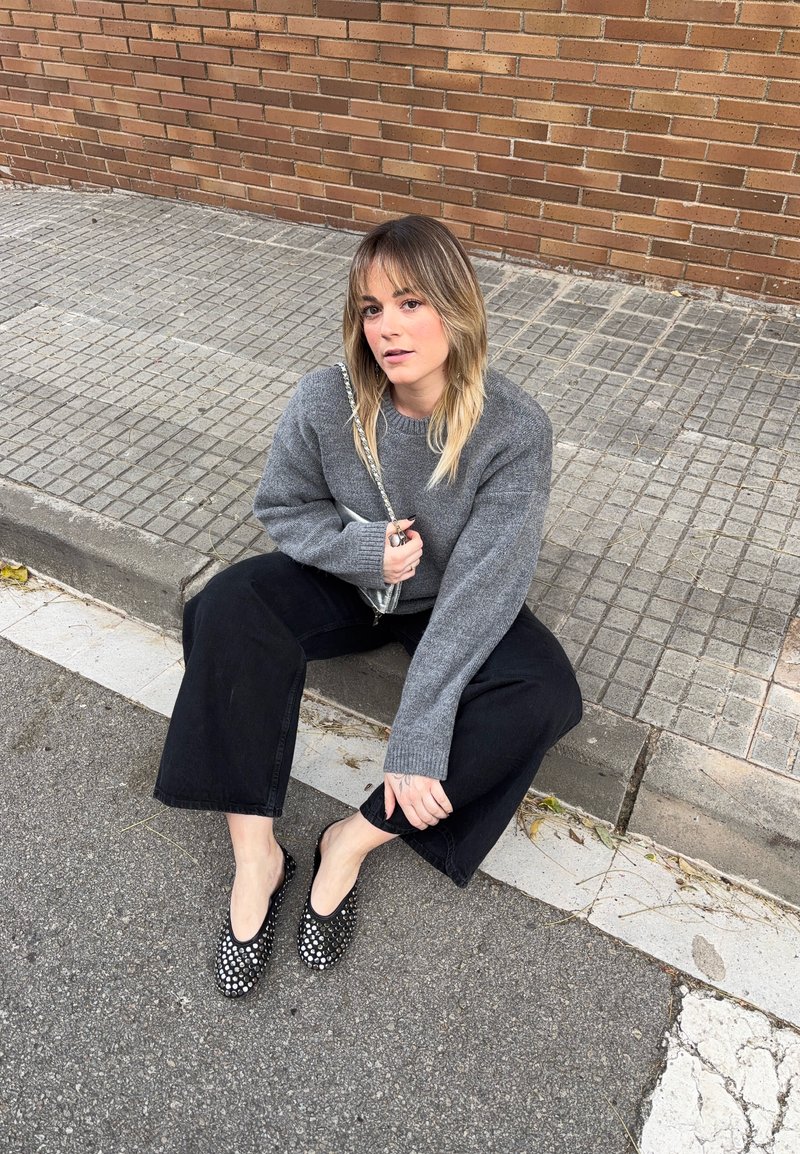Νεαρή γυναίκα με ξανθά μαλλιά μέχρι τους ώμους, καθισμένη σε κράσπεδο, φοράει γκρι πουλόβερ, μαύρο παντελόνι φαρδύ στο κάτω μέρος και μαύρα παπούτσια με τρουκς.