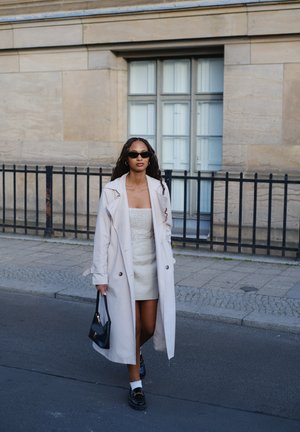 Gabardina beige claro sobre un vestido blanco con textura, gafas de sol negras, bolso negro y zapatos negros con calcetines blancos, de pie en una calle.