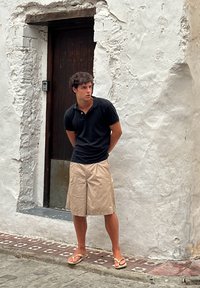 Un tânăr îmbrăcat cu un tricou polo negru și pantaloni scurți bej stă în fața unui zid alb texturat, purtând sandale de plajă, cu o ușă din lemn în spatele său.