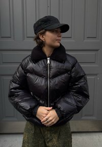 Crna jakna s puf rukavima s teksturiranim ovratnikom od runa i sjajnim završetkom; uparena s crnom kapom. Pozadina prikazuje jednostavna siva vrata.