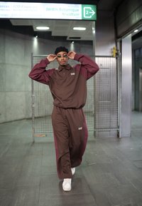 Rjav in maroon trenirka s polovičnim ovratnikom in logotipom, v kombinaciji z belimi supergami. Dodatki vključujejo okrogle sončna očala in črno zimsko kapo.