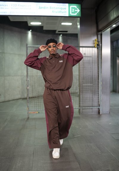 Chándal marrón y burdeos con cuello de polo y logo, combinado con zapatillas blancas. Los accesorios incluyen gafas de sol redondas y un gorro negro.
