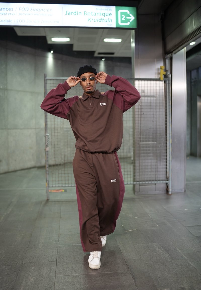 Completo sportivo marrone e bordeaux con colletto a polo e logo, abbinato a sneakers bianche. Gli accessori includono occhiali da sole rotondi e un berretto nero.