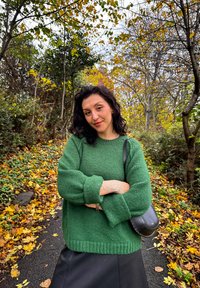 Green knitted sweater with textured fabric and loose sleeves, teamed with a black leather skirt and a small black handbag. Autumn foliage in the background.