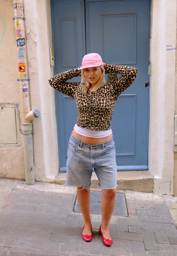 Haut à manches longues en imprimé léopard, short en denim léger bleu déchiré, chapeau seau rose et ballerines rouges. L'arrière-plan présente une porte bleue et un environnement de rue.
