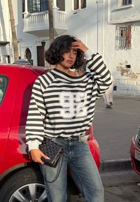 Czarna i biała bluza w szerokie paski z grafiką "92", zestawiona z jasnoniebieskimi dżinsami oraz czarną pikowaną torebką. W tle czerwony samochód.