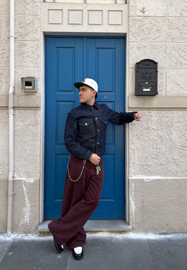 Jeansjacke mit zwei vorderen Taschen, bordeauxrote weite Hose und schwarze Plateauschuhe. Zubehör umfasst eine Kette und Schlüssel. Hintergrund mit blauer Tür.