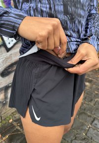 Črne atletske kratke hlače s plisejskim dizajnom, elastičnim pasom in stranskim razporkom. Roka vstavlja ključ v notranji žep.