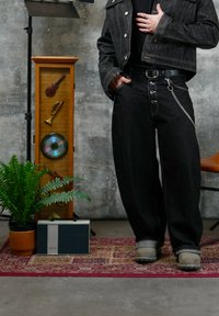 Personne debout en tenue en denim noir, main dans la poche, à côté d'un présentoir musical en bois, d'un fougère en pot, d'une radio vintage et d'un tapis à motifs.