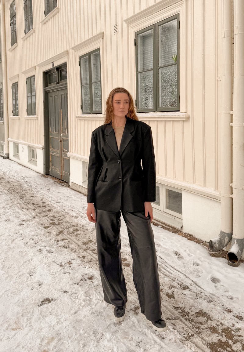 Femme debout dans une rue enneigée à côté d'un bâtiment en bois beige, portant un blazer noir, un pantalon à jambes larges et des baskets, regardant sur le côté.