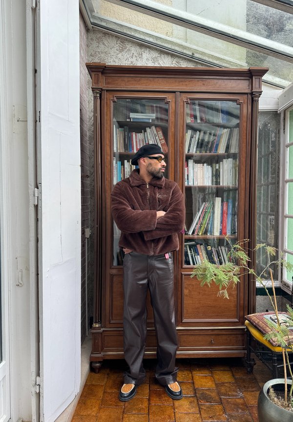 Chaqueta de piel sintética marrón con cuello, pantalones de cuero marrón, boina negra y gafas de sol; de pie frente a una estantería de madera.