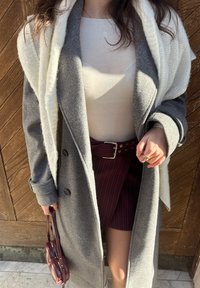 Manteau en laine gris avec de gros boutons, porté par-dessus une chemise blanche. Jupe bordeaux à fines rayures, cintrée à la taille par une ceinture métallique. Petit sac à main rond.