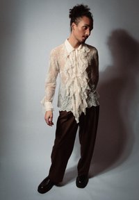 Blouse en dentelle avec des volants en crème, associée à un pantalon marron ample. Présente un col pointu et des poignets boutonnés, portée avec des chaussures noires.