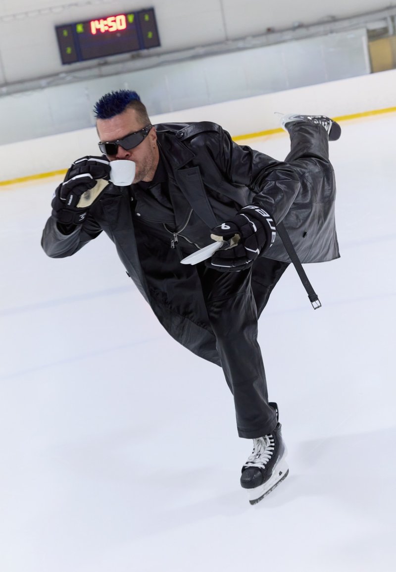 Une patineuse artistique portant un long manteau en cuir noir et des gants noirs glisse sur la glace, tenant une tasse blanche d'une main et un plateau de l'autre.