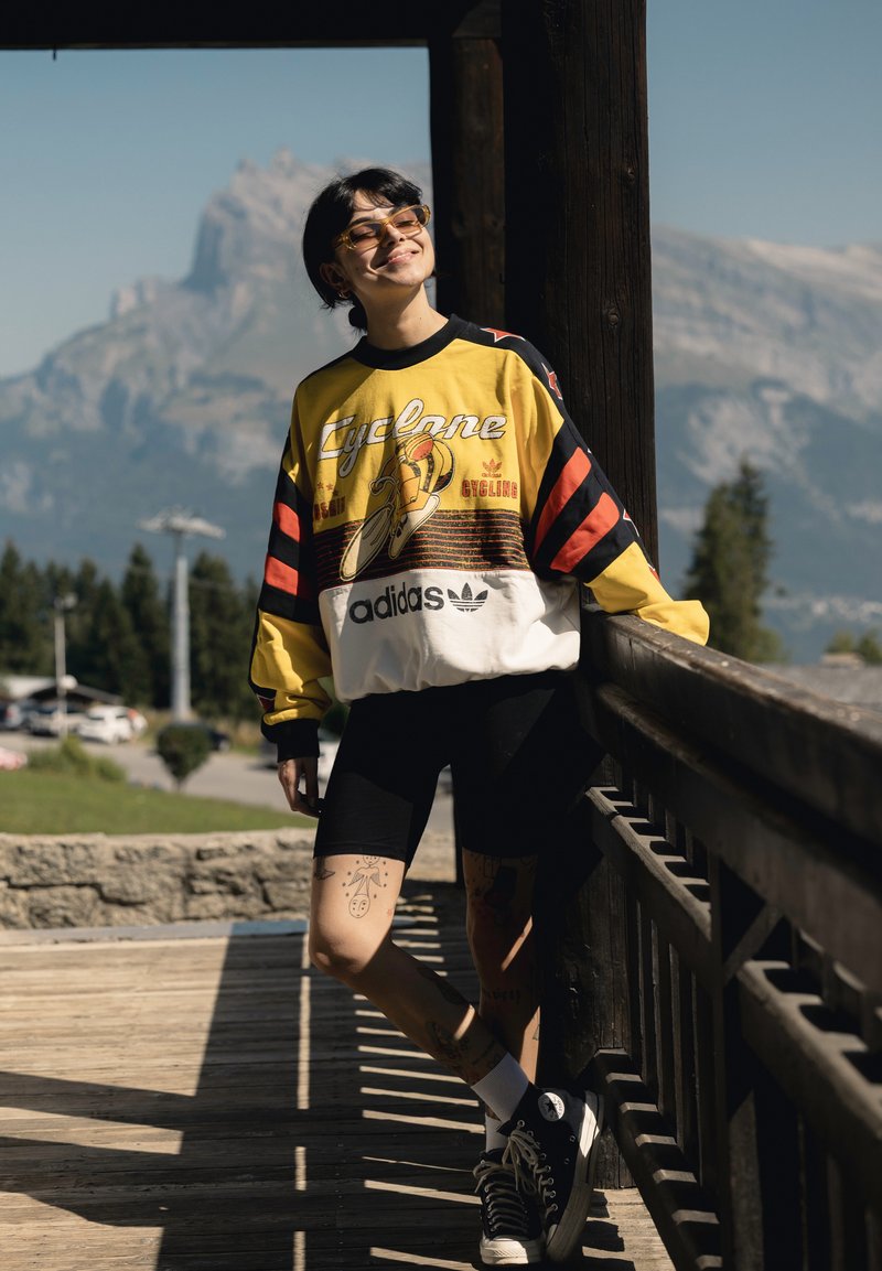 Persona che indossa una felpa da ciclismo Adidas giallo e nero, pantaloncini neri e scarpe da ginnastica, appoggiata a una ringhiera di legno con montagne sullo sfondo.