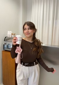 Maglietta marrone a maniche lunghe con fori per il pollice, pantaloni bianchi a vita alta, collana di perle, borsa rosa con un fiore, mentre tiene una tazza di caffè.