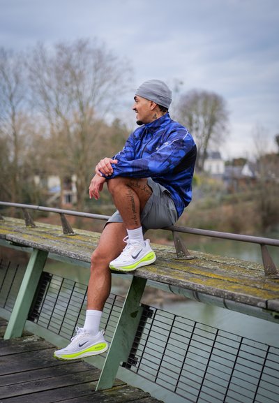 Hombre con chaqueta azul y pantalones cortos grises sentado en una barandilla de madera junto al agua, vestido con zapatillas blancas Nike y un gorro gris, mirando hacia un lado.