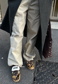 Personne portant un pantalon beige clair et des baskets Adidas à imprimé léopard, tenant un manteau sombre et un sac bordeaux avec des clous argentés, debout sur l'asphalte.