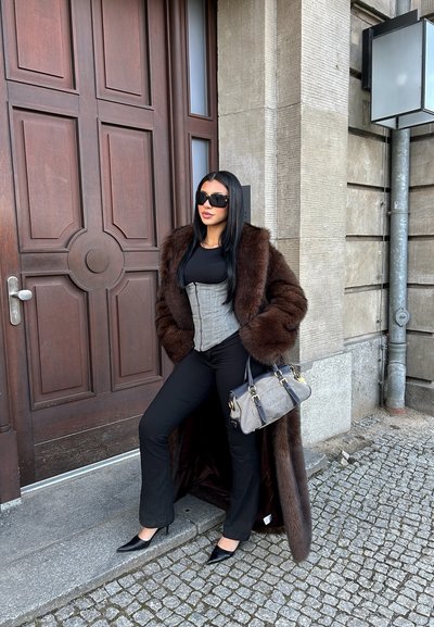 Mujer con gafas de sol oscuras, abrigo de piel marrón, corsé gris, pantalones negros y tacones, sosteniendo un bolso gris de Prada, está junto a una puerta de madera al aire libre.