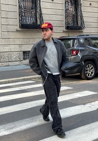 Giacca grigio scuro sopra un maglione grigio chiaro, jeans neri, scarpe nere e un cappello bordeaux con testo giallo. Porta una borsa nera.