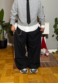 Grauer Strickpullover, weißes Hemd darunter, schwarze weite Hose mit Kettendetail und graue Sneakers, vor einem schlichten Innenraum.