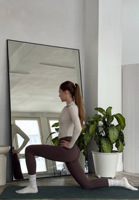 Žena v béžovém topu s dlouhým rukávem a hnědých legínách provádí výpad na zelené podložce, která se odráží v velkém zrcadle s černým rámem. Vedle stojí květináč s rostlinou.