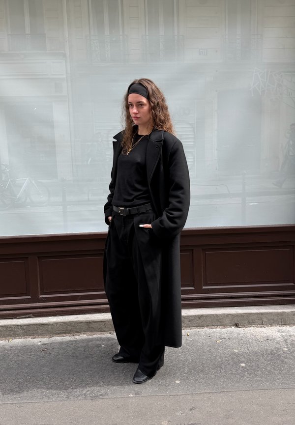 Junge Frau mit langen lockigen Haaren, die ein schwarzes Stirnband, einen langen schwarzen Mantel, schwarze Hose und schwarze Schuhe trägt, steht auf dem Bürgersteig einer Stadt.
