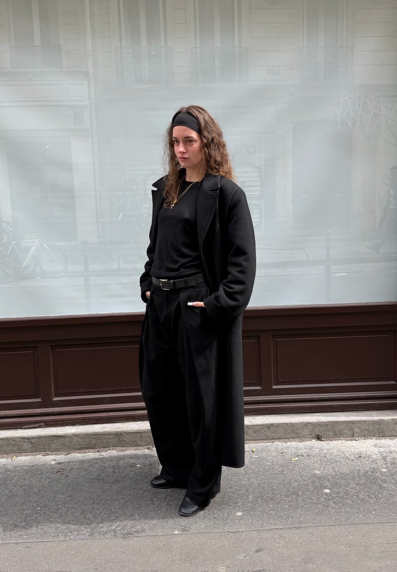 Jeune femme aux longs cheveux bouclés portant un bandeau noir, un long manteau noir, un pantalon noir et des chaussures noires, debout sur un trottoir en ville.
