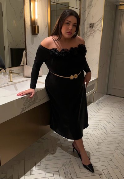 Vestido negro de hombros al descubierto con detalles de volantes florales, ceñido a la cintura con un cinturón dorado que presenta acentos de rosa, combinado con tacones altos negros.
