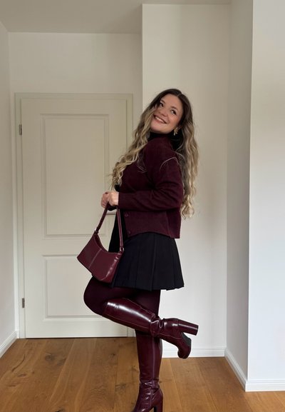 Mujer con cabello largo y ondulado sonríe, lleva un suéter burdeos, botas y bolso a juego, falda negra, posando con una pierna levantada en interiores.