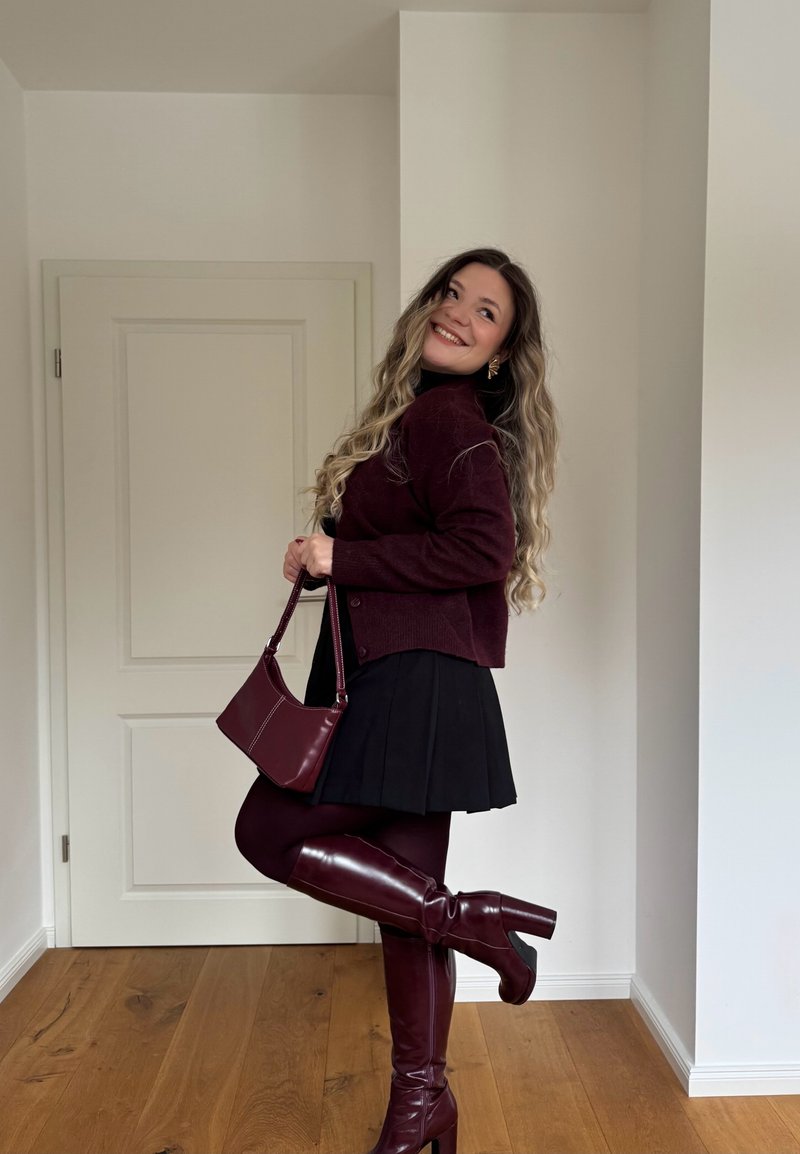 Femme aux longs cheveux ondulés souriant, portant un pull bordeaux, des bottes et un sac assortis, une jupe noire, posant avec une jambe levée à l'intérieur.