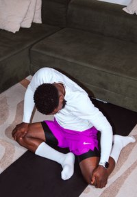 Homme portant une chemise blanche à manches longues et un short violet s'étirant sur un tapis de yoga noir à côté d'un canapé marron dans le salon.