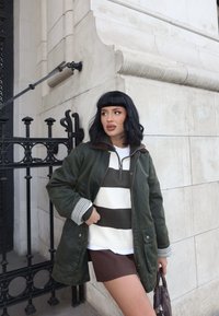 Olivgrüne Jacke mit braunem Kragen, darunter ein weiß-grün gestreiftes Oberteil und braune Shorts. Als Accessoires dienen eine Handtasche und kariertes Bündchen.