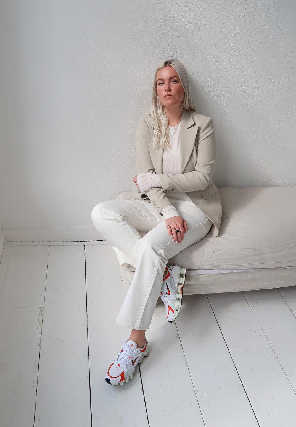 Lichtbeige op maat gemaakte blazer over een witte top, gecombineerd met witte uitlopende broek en witte sneakers met rode accenten, zittend op een lichte bank.