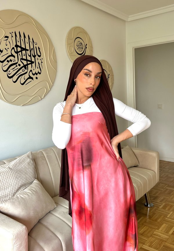 Femme portant un hijab marron et une robe rose posant avec une main sur son cou et l'autre sur sa hanche dans un salon.