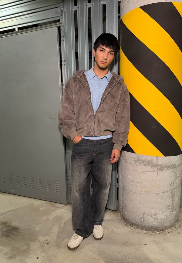 Jeune homme en veste grise et jean se tient avec la main dans la poche, appuyé contre un pilier en béton avec des bandes noires et jaunes.