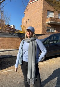 Jeune femme portant une casquette bleue, un pull bleu clair, une écharpe grise et un pantalon noir, debout dans une rue ensoleillée avec des bâtiments en briques et des voitures à proximité.