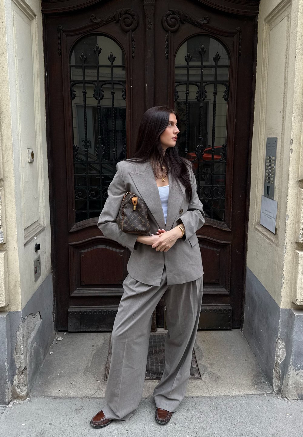 Costum gri supradimensionat cu rever ascuțite, pantaloni largi și un top alb. Accesorizat cu o poșetă maro cu un model și pantofi maro.