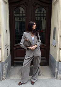 Costum gri supradimensionat cu rever ascuțite, pantaloni largi și un top alb. Accesorizat cu o poșetă maro cu un model și pantofi maro.