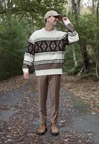 A man adjusting his cap, dressed in a patterned sweater, brown trousers, and boots, stands on a leaf-strewn path in a wooded outdoor setting.