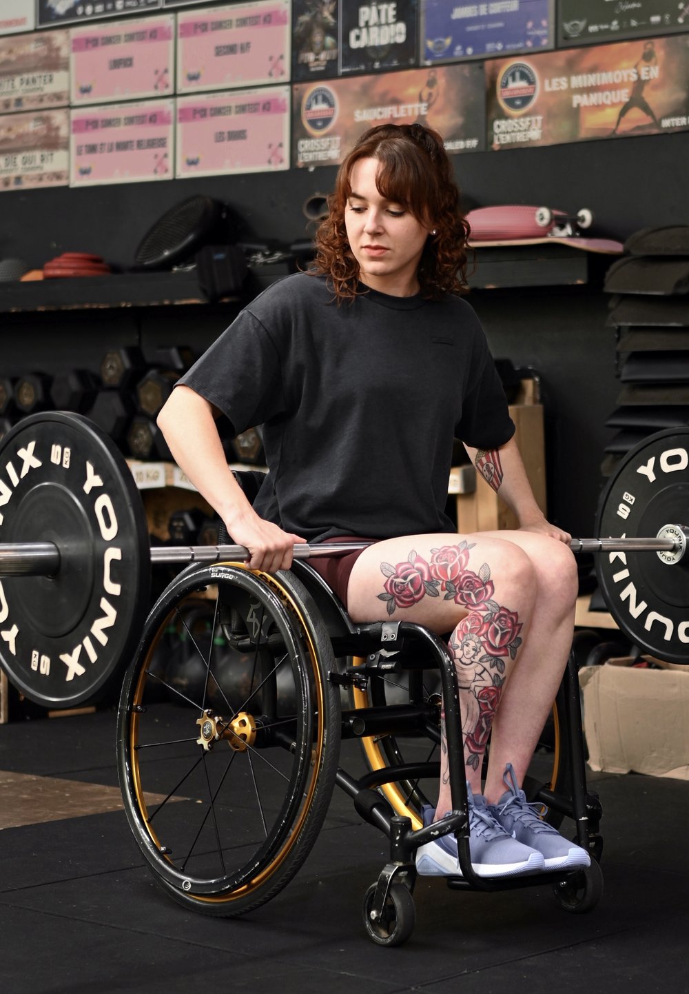 Atleta com uma camisola preta e calções bordô, sentado numa cadeira de rodas preta e dourada, segurando um haltere, com tatuagens florais nas pernas.