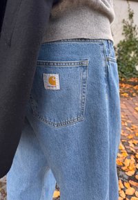 Šviesiai mėlynos spalvos Džinsai iš džinso audinio su laisvu kirpimu, turintys kišenę gale su geltonu "Carhartt" logotipo ženkleliu, matomais siūlais ir tekstūra.