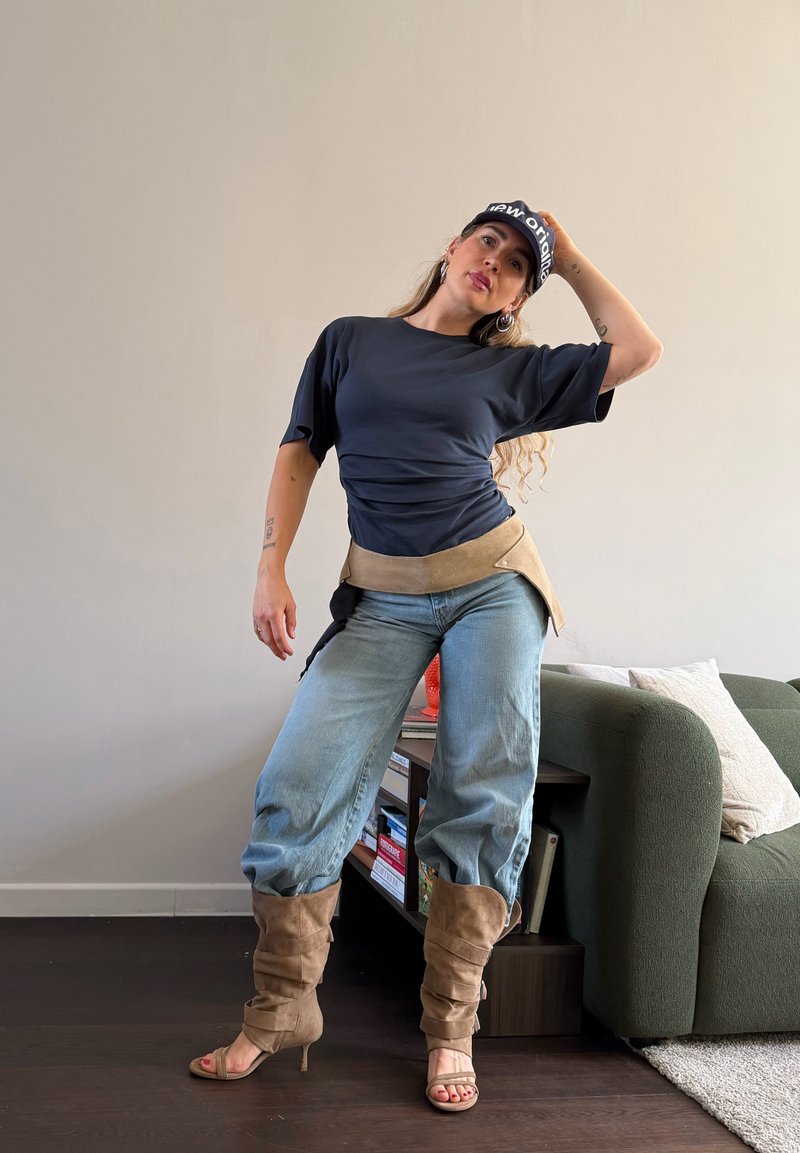 Femme en tenue décontractée et bottes pose à l'intérieur près d'une bibliothèque et d'un canapé vert, portant une casquette, une chemise foncée, un jean et des accessoires beige.